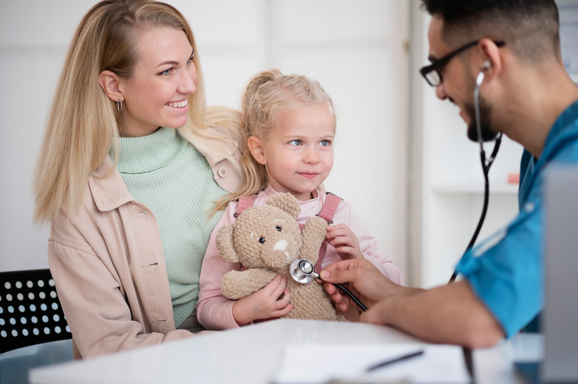 A mother and her daughter with a provider listening to a teddy bear's heartbeat